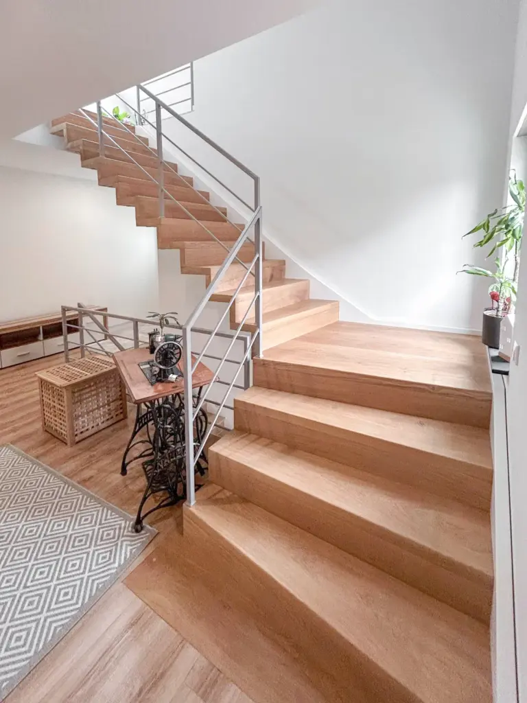 Eine moderne Innentreppe mit hellen Holzstufen und Metallgeländer, ein gemusterter Teppich, ein kleiner Tisch mit einer alten Nähmaschine und eine Topfpflanze an einem Fenster auf der rechten Seite.