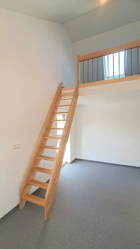Ein minimalistischer Raum mit grauem Teppich, weißen Wänden, einer Holztreppe, die zu einem Loft mit Geländer führt, und einem großen Fenster, durch das Tageslicht in den oberen Bereich fällt.
