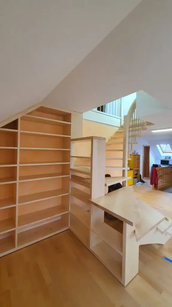Unter einer Treppe in einer hellen, modernen Wohnung mit hellem Holzfußboden und offener Raumaufteilung sind maßgefertigte Holzregale und ein kleiner Schreibtisch angebracht.