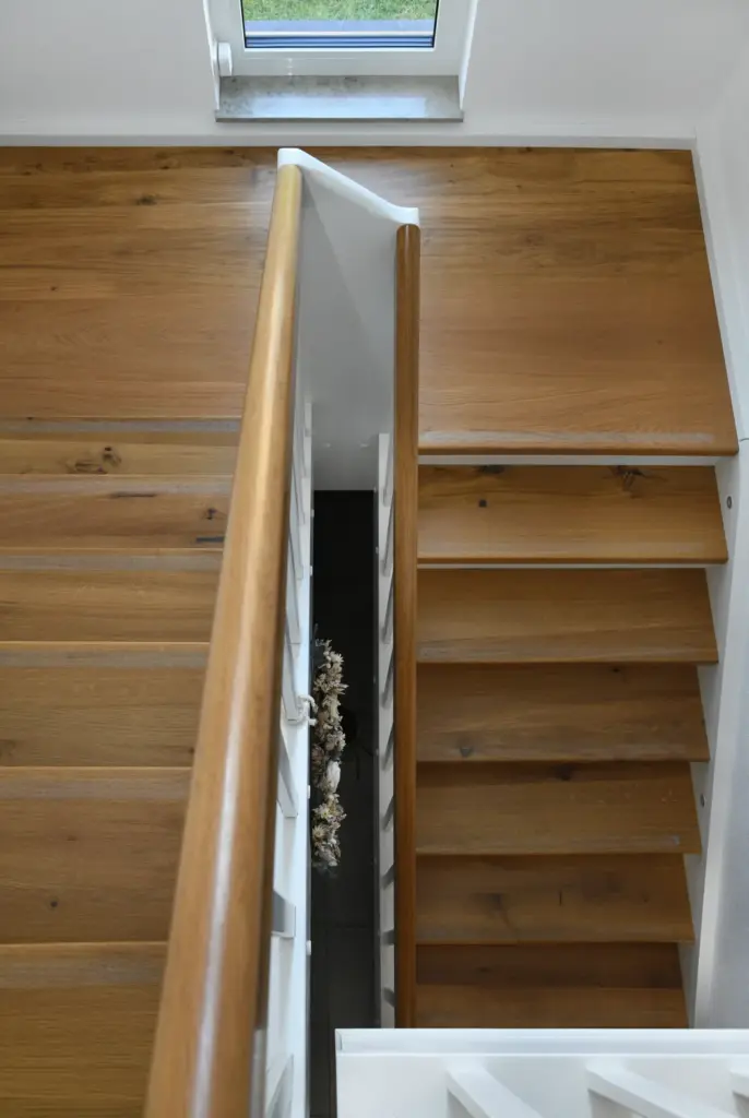 Draufsicht auf eine Holztreppe mit hellbraunem Handlauf, weißem Geländer und einem kleinen Fenster am oberen Treppenabsatz. Die Treppe führt zu einer unteren Etage mit dunklem Bodenbelag und einigen sichtbaren Dekorationsgegenständen.