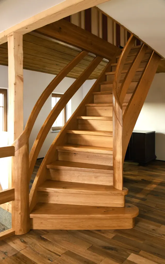 Eine Holztreppe mit geschwungenen Handläufen und breiten Stufen in einem Raum mit Holzböden, sichtbaren Balken und großen Fenstern, die viel Tageslicht hereinlassen.