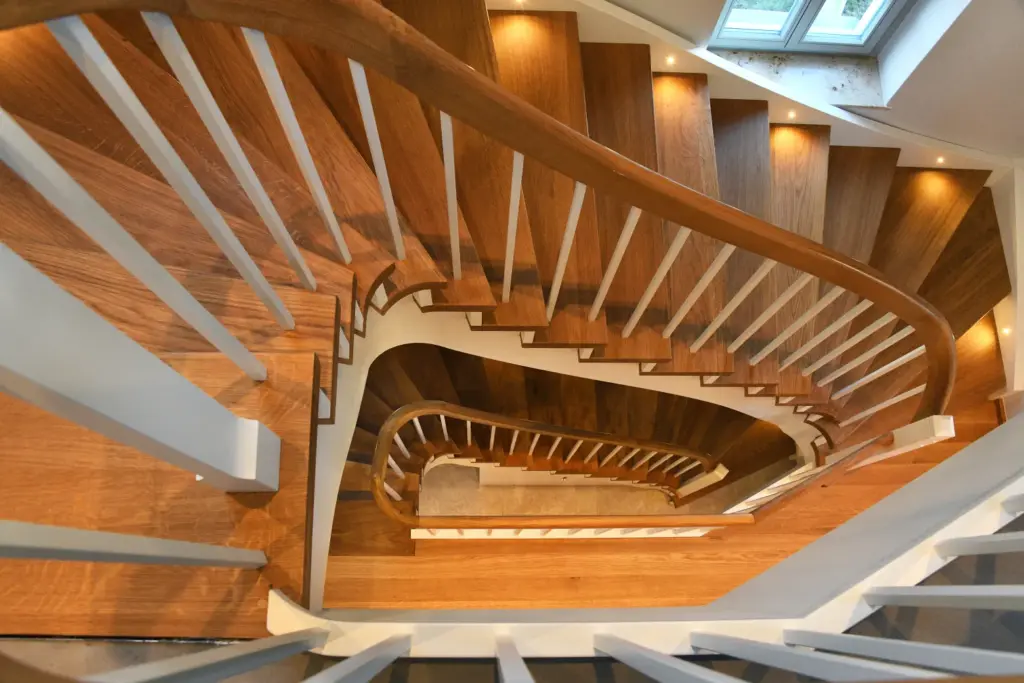 Eine Wendeltreppe mit Holzstufen, weißem Geländer und geschwungenen Holzhandläufen, von oben gesehen. Kleine Lichter sind entlang der Treppe in die Wand eingelassen. Am oberen Rand des Bildes ist ein Fenster zu sehen.