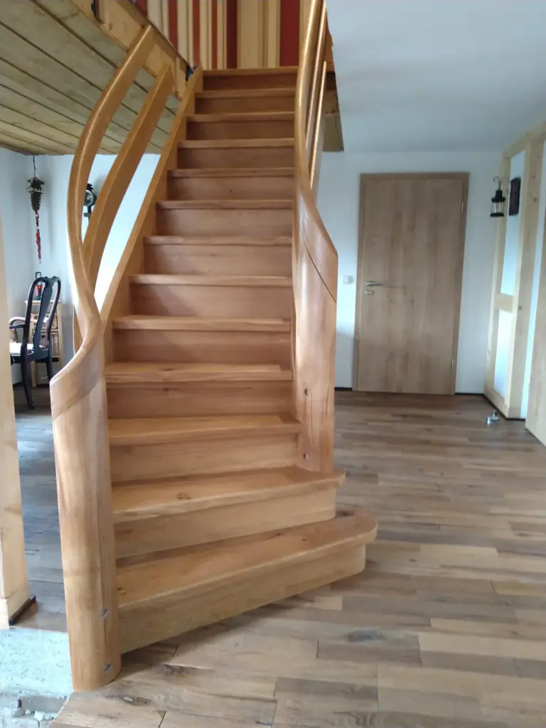 Eine Holztreppe mit geschwungenen Handläufen führt in einem Haus mit Holzfußboden und hellen Wänden nach oben. Im Hintergrund sind eine geschlossene Holztür und ein Teil eines Essbereichs zu sehen.