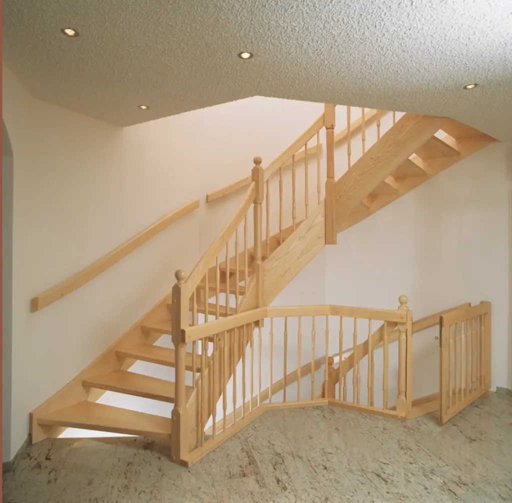 Eine helle Holztreppe mit offenen Setzstufen und passendem Geländer schlängelt sich in einem hellen Innenraum nach oben. Am Fuß der Treppe ist ein hölzernes Sicherheitstor auf einem Steinboden installiert. Darüber sind Deckeneinbauleuchten zu sehen.
