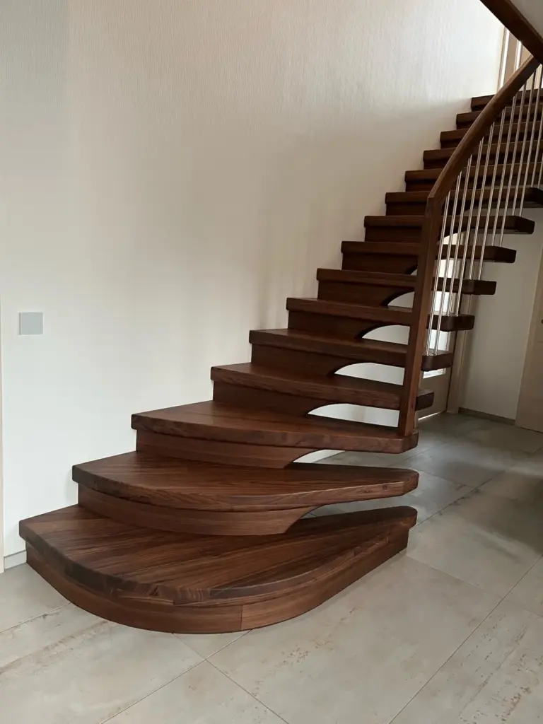 Eine moderne, geschwungene Holztreppe mit offenen Setzstufen führt neben einer weißen Wand nach oben. Die Stufen aus dunklem Holz sind dick und einzigartig geformt, so dass ein schwebender Effekt entsteht. Der Boden besteht aus hellen Fliesen.