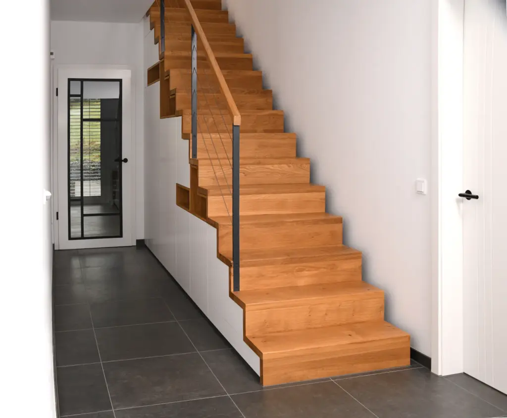 Ein moderner Flur mit großen grauen Bodenfliesen und einer Holztreppe, unter der ein Stauraum eingebaut ist. Weiße Wände und Türen sowie eine Glastür am Ende des Flurs lassen natürliches Licht herein.