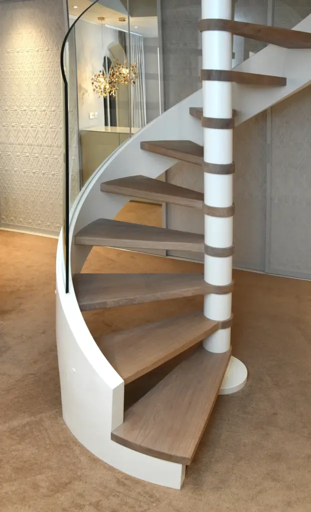 Eine moderne Wendeltreppe mit Holzstufen, einer weißen Mittelsäule und einem geschwungenen Glasgeländer vor einem beigen Teppichboden und hellen, strukturierten Wänden.