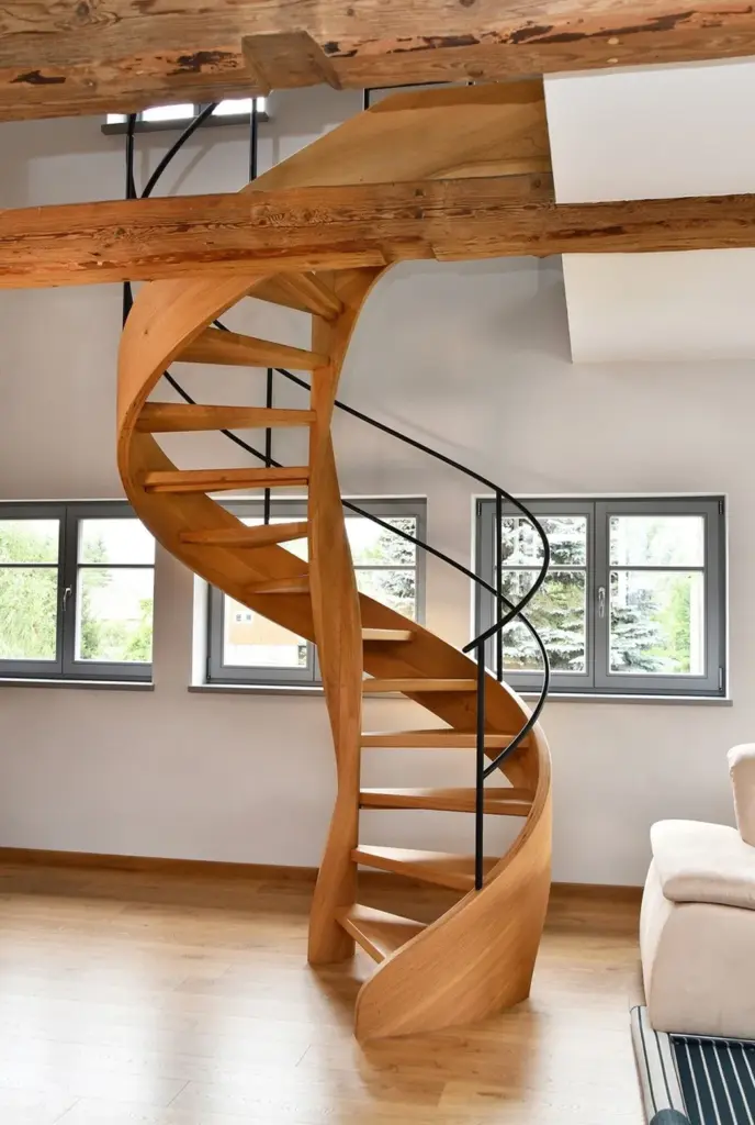 Eine moderne Holzspindeltreppe mit offenen Stufen und schwarzem Handlauf steht in einem hellen Raum mit hellen Wänden, großen Fenstern und freiliegenden Holzbalken an der Decke.