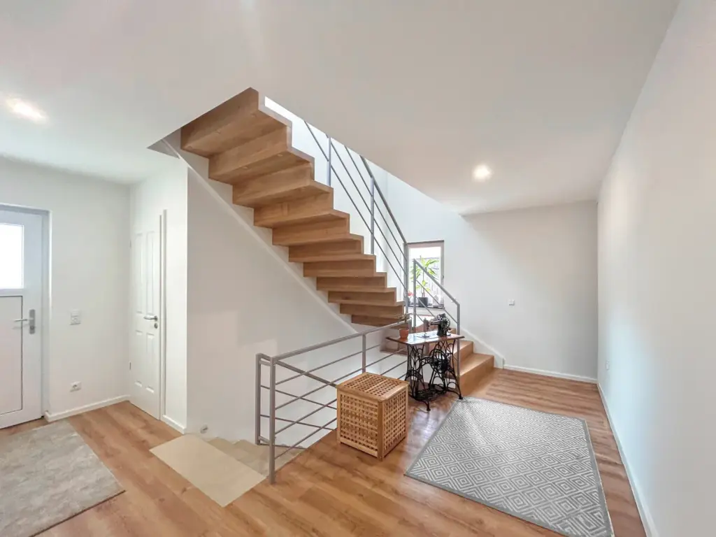 Ein heller, moderner Flur mit Holzfußboden, einer Treppe mit Holzstufen und einem Metallgeländer, einem geflochtenen Korb und einem Schreibtisch mit einer Pflanze in der Nähe eines großen Fensters. Ein grau gemusterter Teppich liegt auf dem Boden.