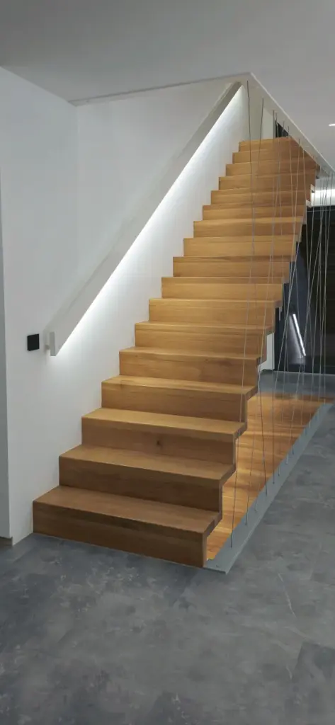 Eine moderne Holztreppe mit offenen Setzstufen, schlankem Kabelgeländer und eingebauter LED-Beleuchtung entlang der weißen Wand, die von einem grauen Fliesenboden in einem zeitgenössischen Interieur nach oben führt.