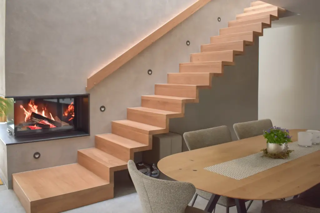 Ein moderner Essbereich mit Holztisch und grauen Stühlen, daneben eine schwebende Holztreppe und ein eingebauter Kamin in einer minimalistischen Betonwand.