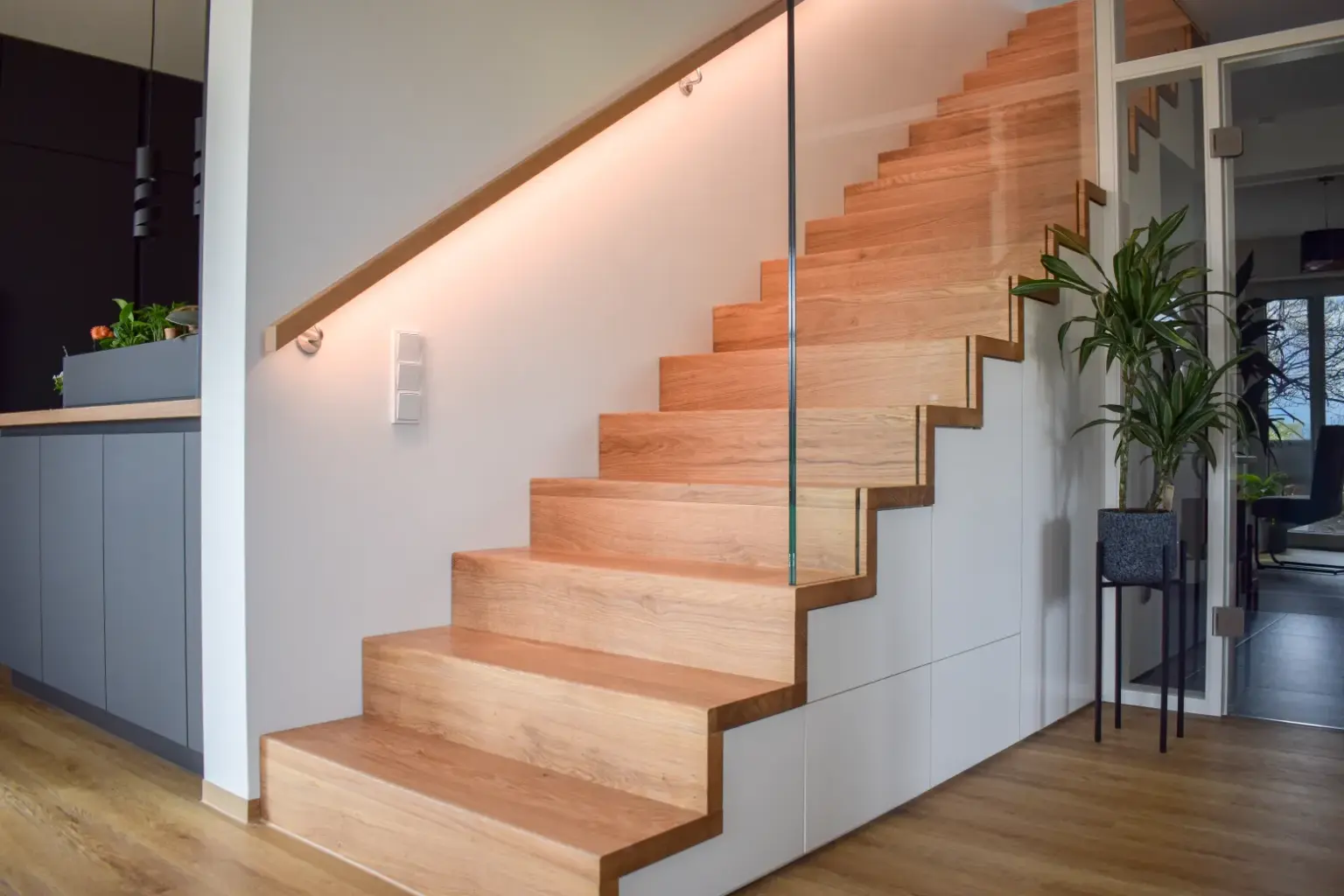 Eine moderne Holztreppe mit Glasgeländer und eingebauter Beleuchtung, daneben eine grüne Topfpflanze und ein Küchenbereich mit dunklen Schränken in einem hellen, zeitgenössischen Wohnbereich.