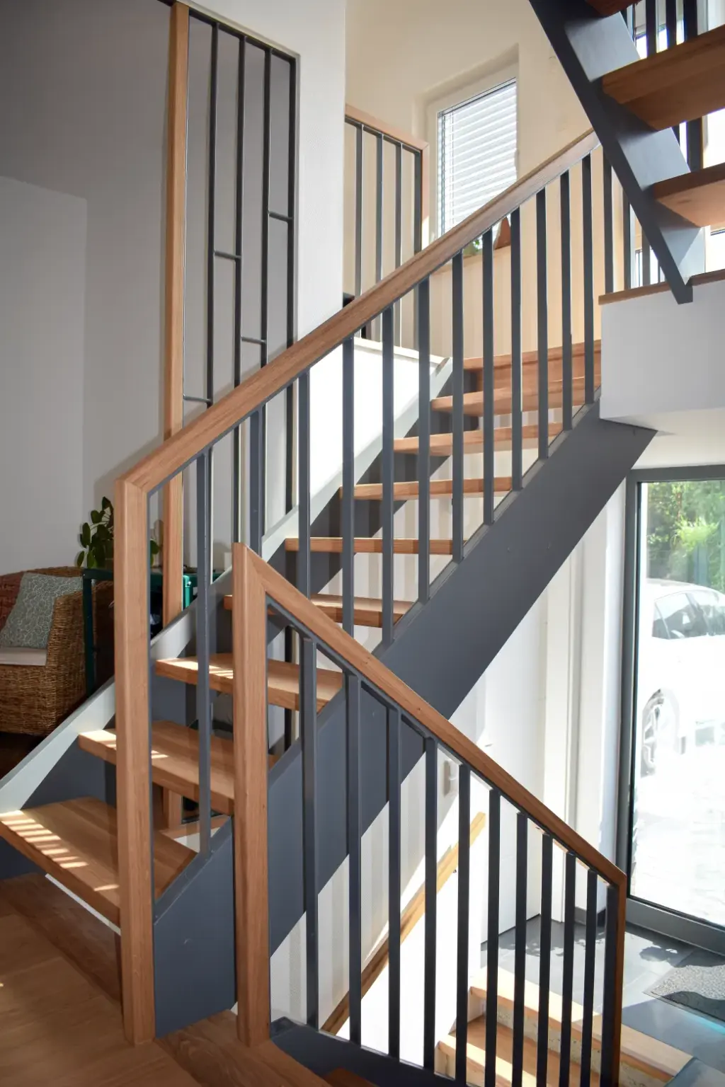 Eine moderne Holztreppe mit schwarzem Metallgeländer, die mehrere Läufe und einen Treppenabsatz aufweist. Sonnenlicht strömt aus den nahe gelegenen Fenstern herein und beleuchtet die Treppe und Teile eines Wohnraums mit einem Korbstuhl.