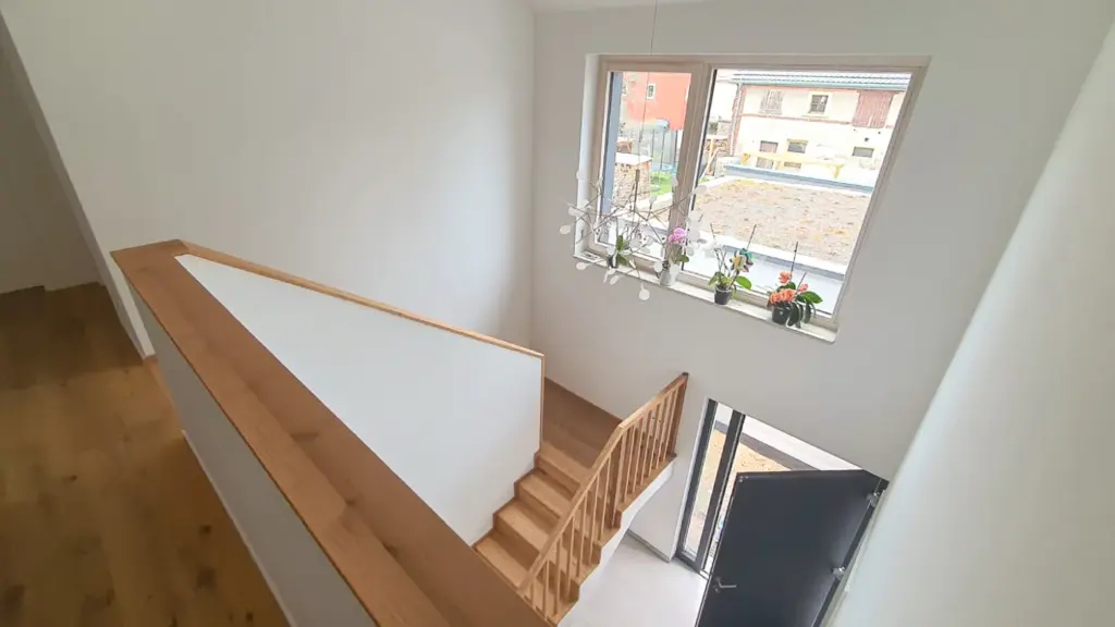 Blick von einem Flur im Obergeschoss auf eine Holztreppe mit einem modernen Kronleuchter, einem großen Fenster mit Topfpflanzen und einer offenen Eingangstür, durch die Tageslicht einfällt.