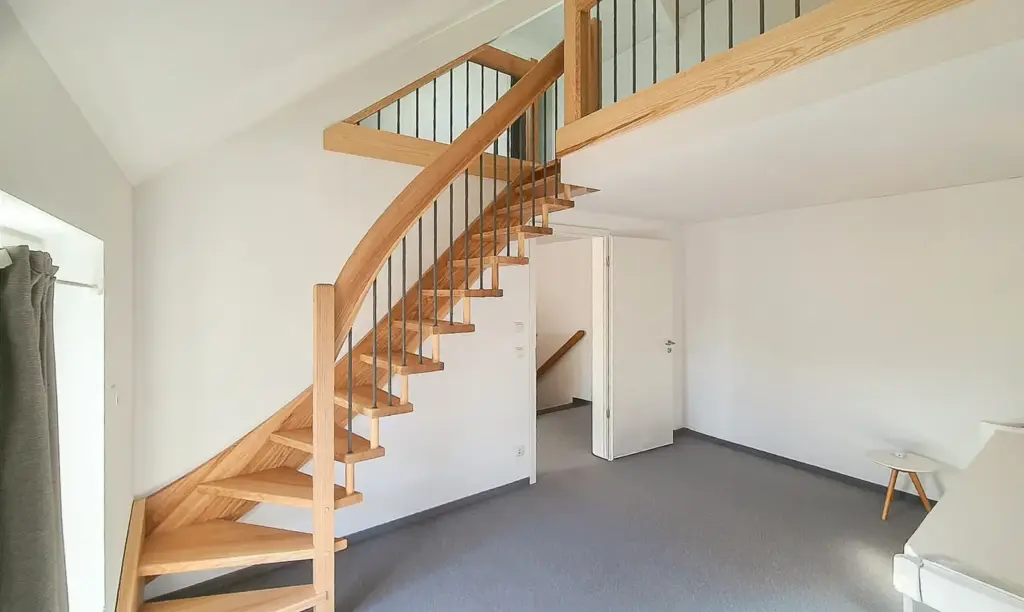 Ein heller, moderner Raum mit grauem Teppich, weißen Wänden und einer hölzernen Wendeltreppe, die zu einem Loftbereich führt. Ein kleiner runder weißer Tisch und ein Sofa sind teilweise auf der rechten Seite zu sehen. Natürliches Licht fällt durch ein Fenster auf der linken Seite ein.