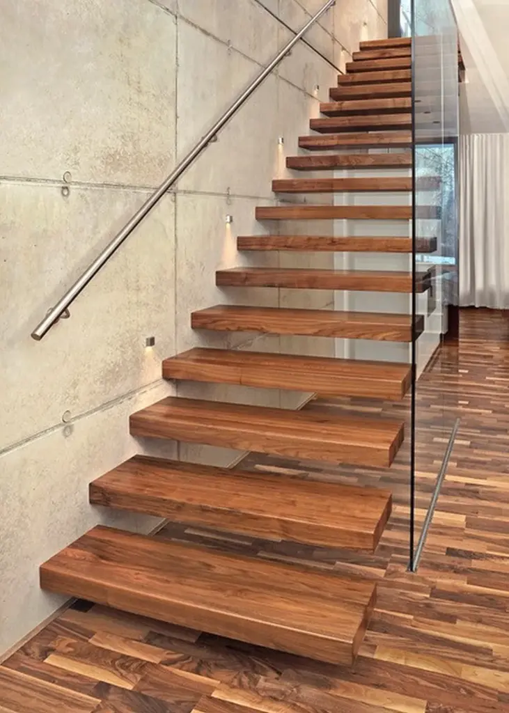 Eine moderne schwebende Holztreppe mit dicken Holzstufen, Glaswange, Metallhandlauf und minimalistischer Wandbeleuchtung vor einer Betonwand und einem Holzboden.