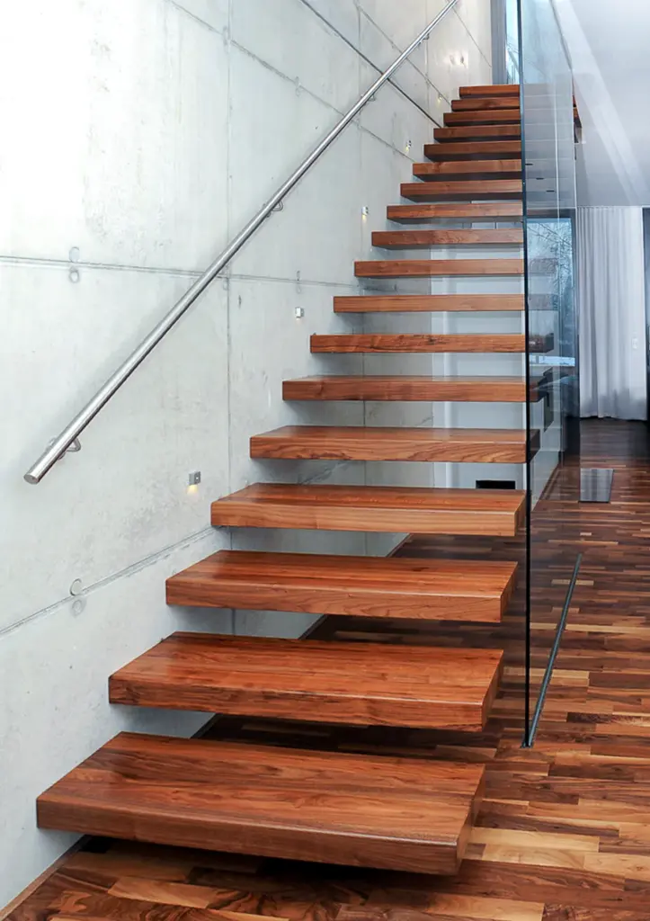 Eine moderne schwebende Holztreppe mit dicken, an einer Betonwand befestigten Stufen, einem Metallhandlauf und einer Seitenwand aus Glas, die von einem Holzboden in einem zeitgenössischen Innenraum nach oben führt.