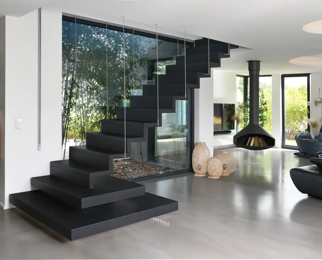 Modernes Wohnzimmer mit eleganter schwarzer Schwebetreppe, Glasgeländer, poliertem Betonboden, großen Fenstern, schwarzem Hängekamin und natürlichen Dekoelementen wie geflochtenen Körben und Zimmerpflanzen.