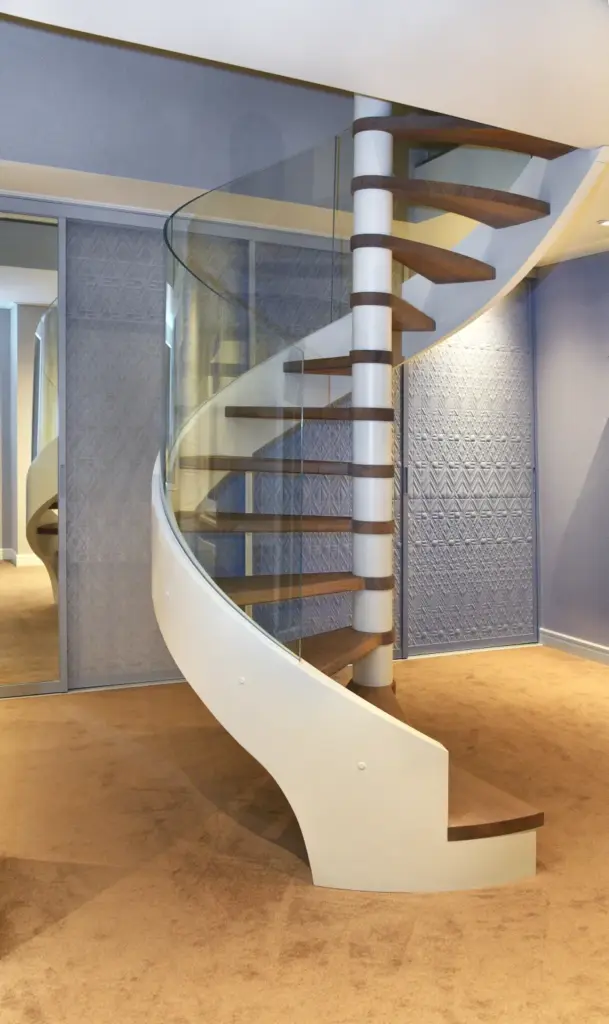 Eine moderne Wendeltreppe mit Holzstufen und einem Glasgeländer vor einer strukturierten blauen Wand und einem beigen Teppichboden in einem modernen Innenraum.