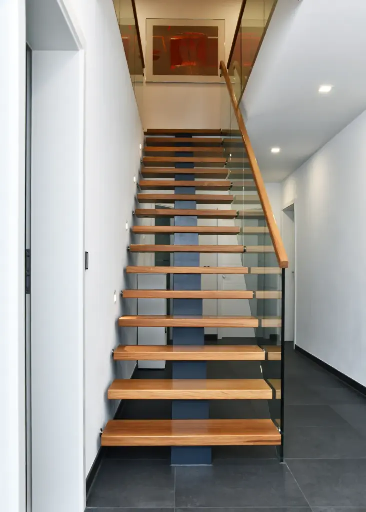 In einem hellen, minimalistischen Flur mit weißen Wänden und dunkelgrauen Bodenfliesen führt eine moderne Treppe mit Holzstufen, Glasgeländer und einer dunklen Mittelstütze nach oben. An der Wand darüber hängt ein abstraktes Kunstwerk.