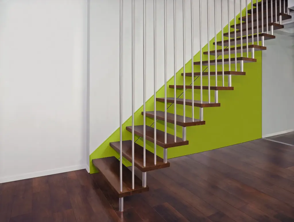 Moderne Treppe mit dunklen Holzstufen, schlanken vertikalen Metallgeländern und einer hellgrünen Wand darunter, die sich von einer weißen Wand und einem dunklen Holzfußboden abhebt.