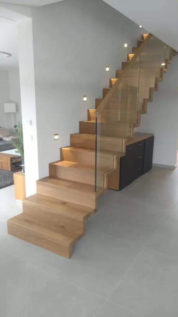 Eine moderne schwebende Holztreppe mit Glasgeländer und eingebauten Wandleuchten führt entlang einer weißen Wand in einem minimalistischen Wohnraum mit hellgrauen Böden nach oben.
