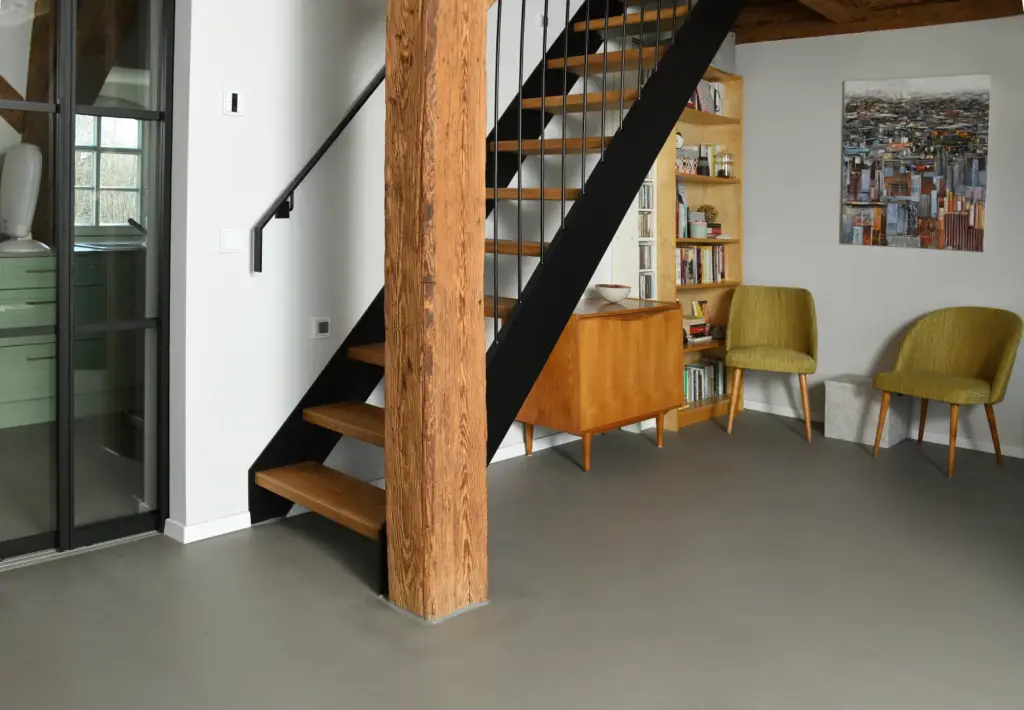 Modernes Interieur mit einer Holztreppe, schwarzem Geländer, freiliegenden Holzbalken, zwei grünen Stühlen, einem Holzschrank, einem Bücherregal und einem Gemälde mit Stadtansichten an einer weißen Wand. Hellgrauer Boden und großes Fenster sichtbar.