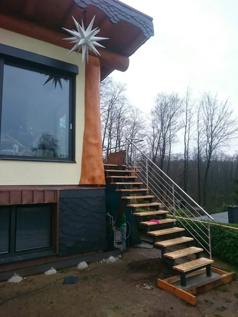 Eine Holzaußentreppe mit Metallgeländer führt zu einem Haus mit einem großen Fenster hinauf. Vom Dach hängt eine sternförmige Dekoration herab. Im Hintergrund sind Bäume ohne Blätter zu sehen.