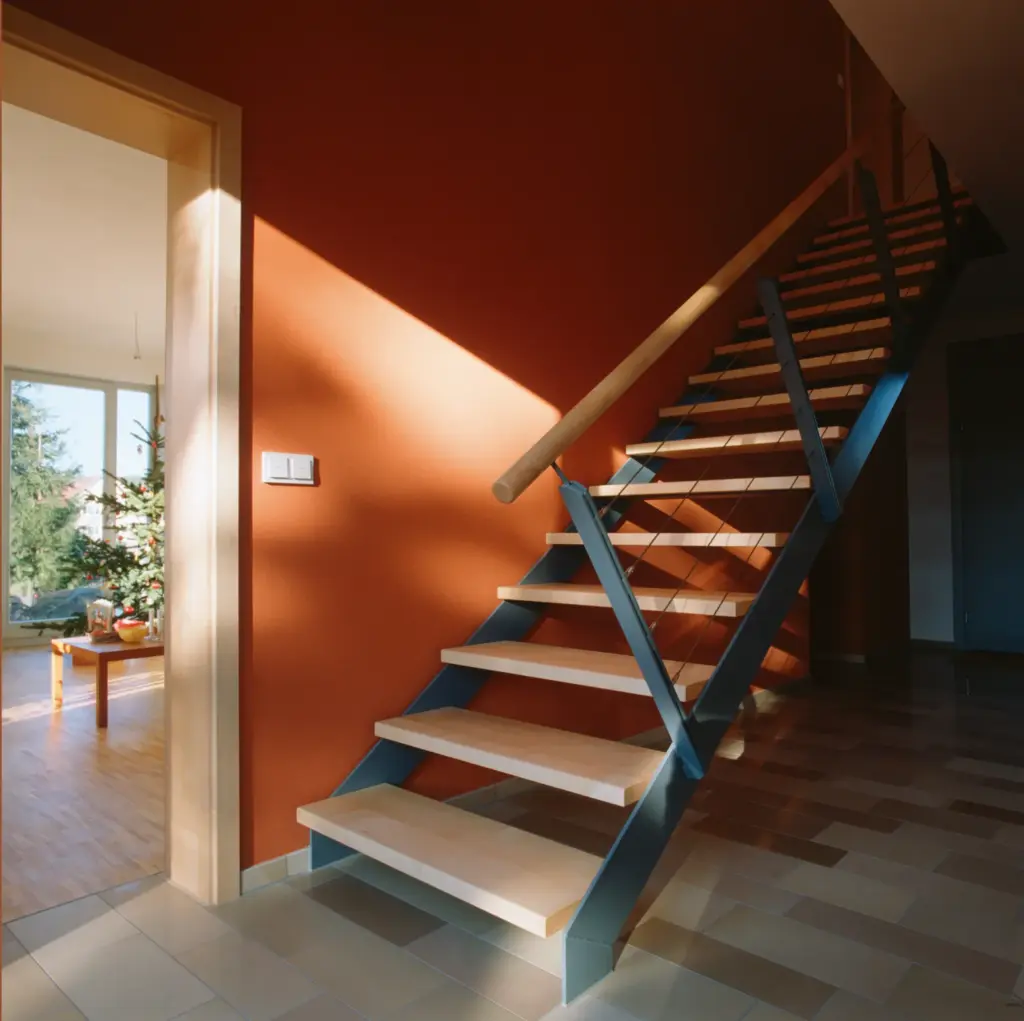 Eine moderne Treppe mit Holzstufen und Metallgeländer steht vor einer orangefarbenen Wand. Das Sonnenlicht fällt durch ein nahe gelegenes Fenster und wirft Schatten auf den gefliesten Boden. In einem angrenzenden Raum ist ein geschmückter Weihnachtsbaum zu sehen.