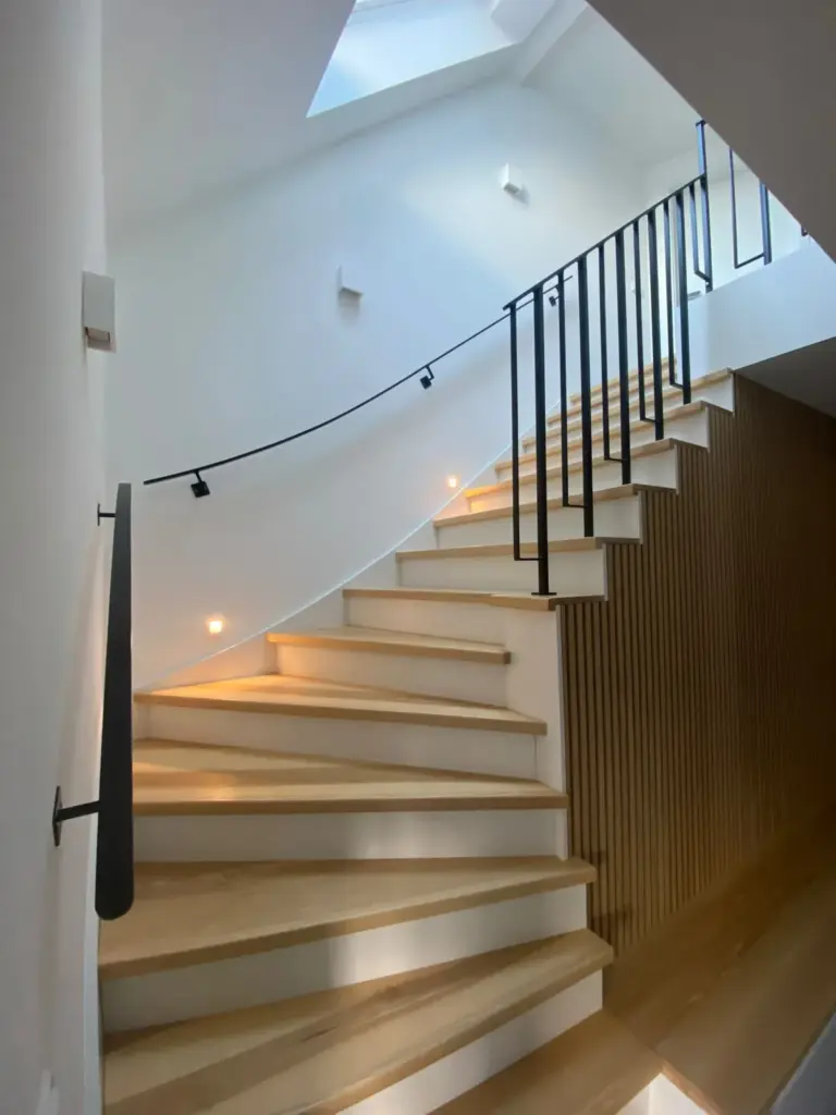 Eine moderne Holztreppe mit schwarzen Handläufen schlängelt sich nach oben und wird von kleinen Lampen entlang der Stufen beleuchtet. Durch ein Oberlicht über der Treppe fällt natürliches Licht ein, das die helle, minimalistische Inneneinrichtung unterstreicht.