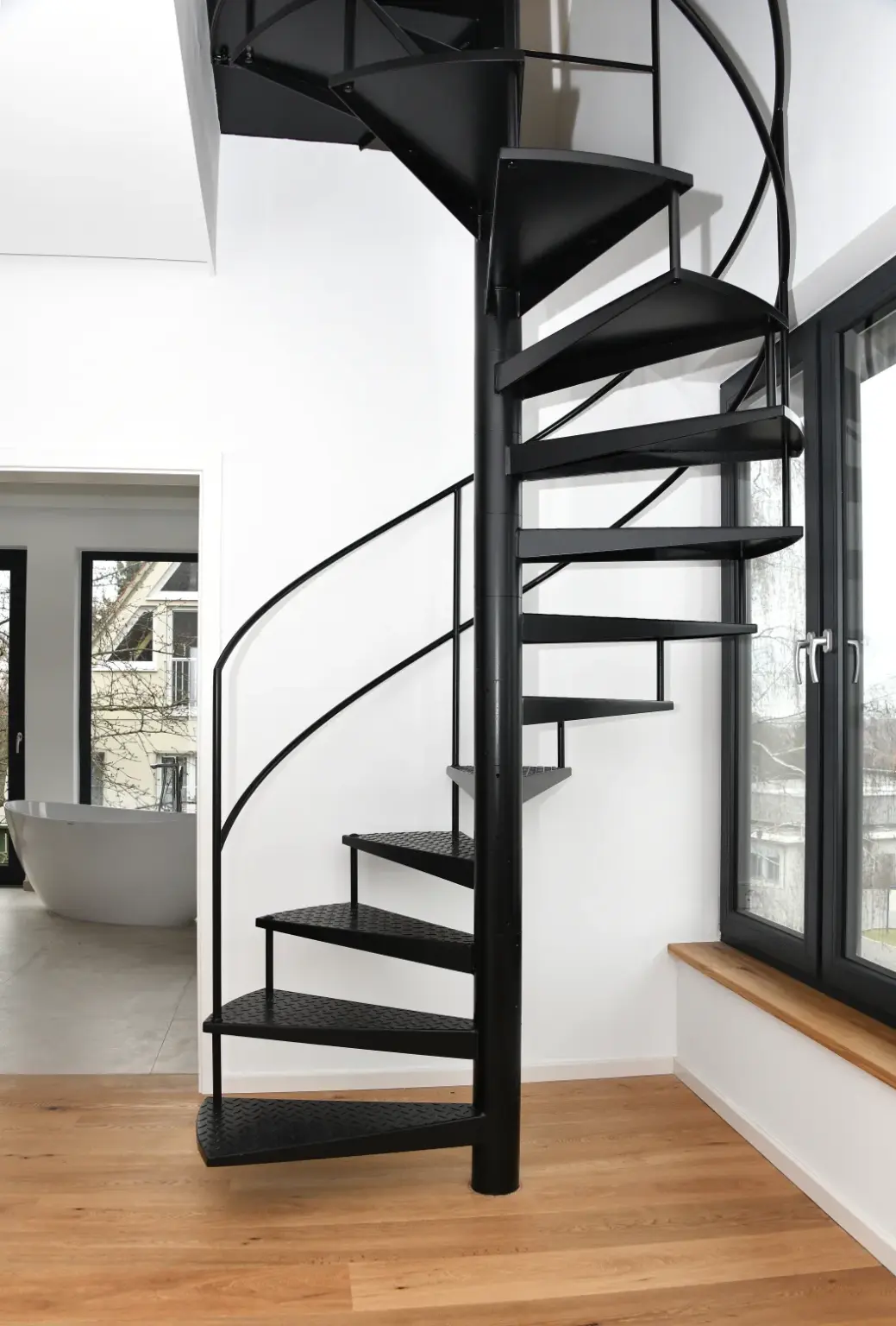Eine moderne schwarze Wendeltreppe mit Metallstufen steht im Innenbereich neben großen Fenstern und einem Holzfußboden und führt zu einem oberen Stockwerk in einem hellen, minimalistischen Raum.