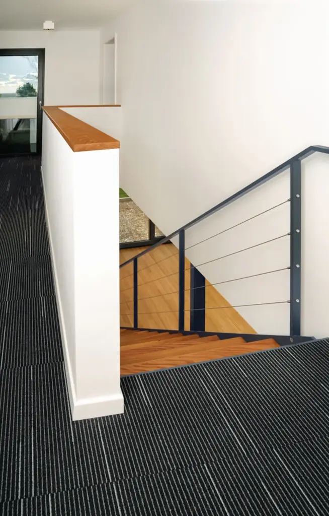 Eine moderne Treppe mit Holzstufen und Metallgeländer führt von einem Flur mit dunklem, gestreiftem Teppichboden und weißen Wänden hinunter; durch ein Fenster am Ende fällt Sonnenlicht ein.
