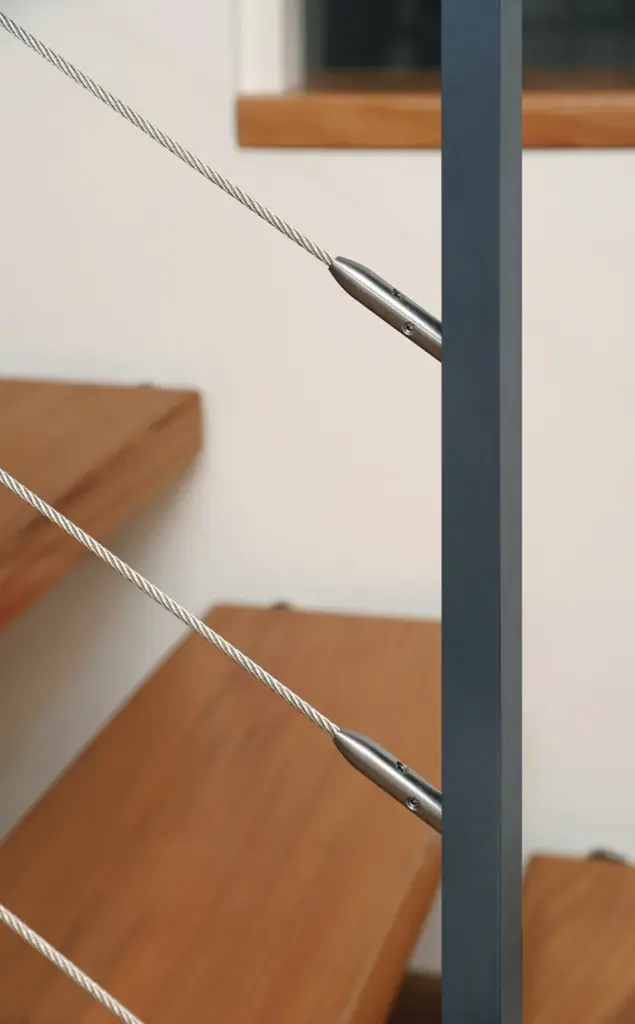 Nahaufnahme einer modernen Treppe mit Holzstufen und einem mit Metallbeschlägen befestigten Metallseilgeländer, das an einem vertikalen dunklen Metallpfosten befestigt ist.