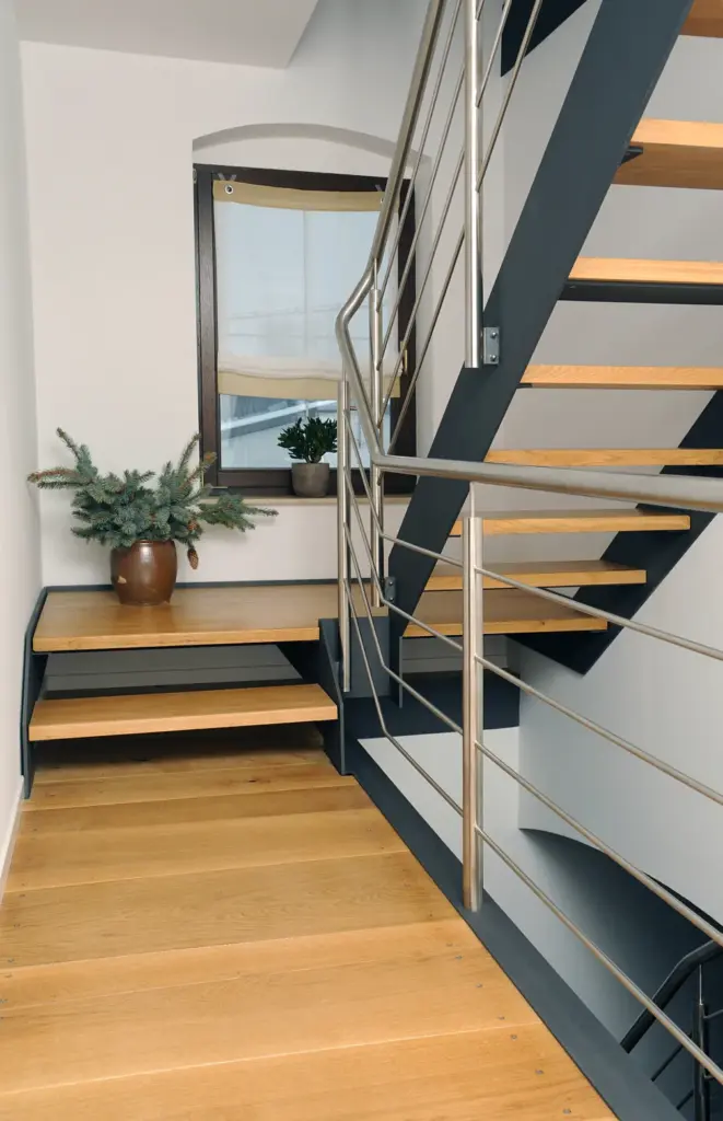 Moderne Innentreppe mit Holzstufen und Metallgeländer, mit einer Topfpflanze auf dem Treppenabsatz neben einem Fenster mit Holzrahmen und beigem Rollo.