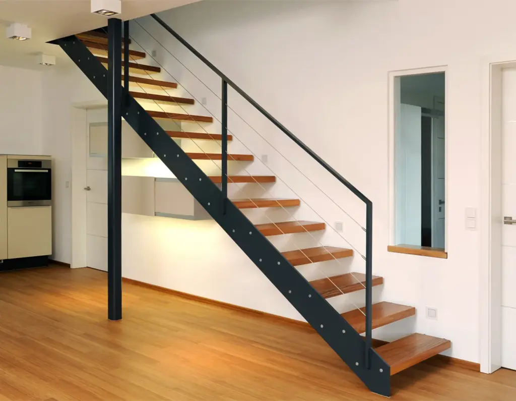 Moderne Einrichtung mit einer minimalistischen Holztreppe mit schwarzem Metallgeländer vor weißen Wänden und hellen Holzböden. Die Treppe führt in ein Obergeschoss, in dem auf der linken Seite eine kleine Küche zu sehen ist.