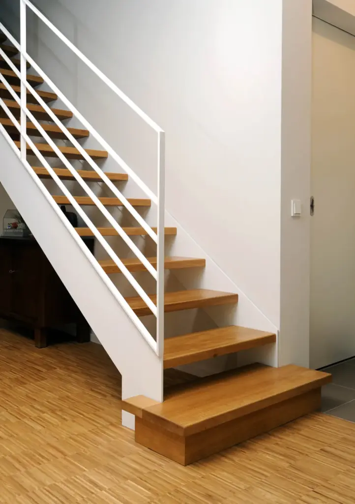 Eine moderne Holztreppe mit offenen Setzstufen und weißem Metallgeländer vor einer weißen Wand in einem Raum mit hellem Holzfußboden.