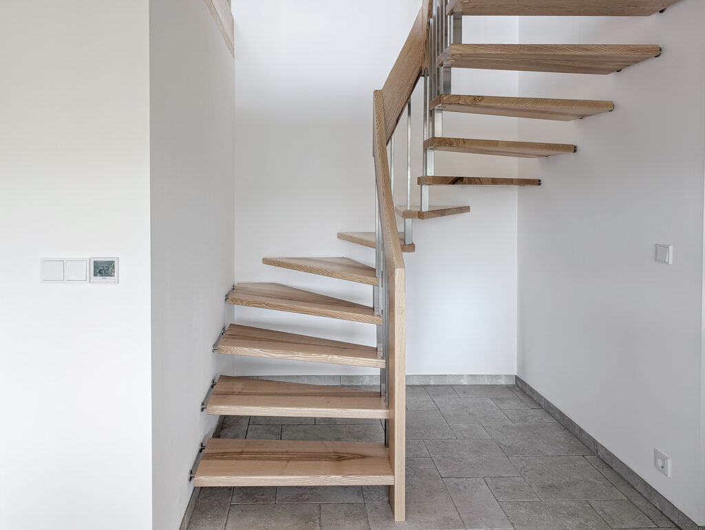 Eine moderne schwebende Holztreppe mit offenen Setzstufen und Metallgeländer in einem hellen, minimalistischen Innenraum mit gefliestem Boden und weißen Wänden.