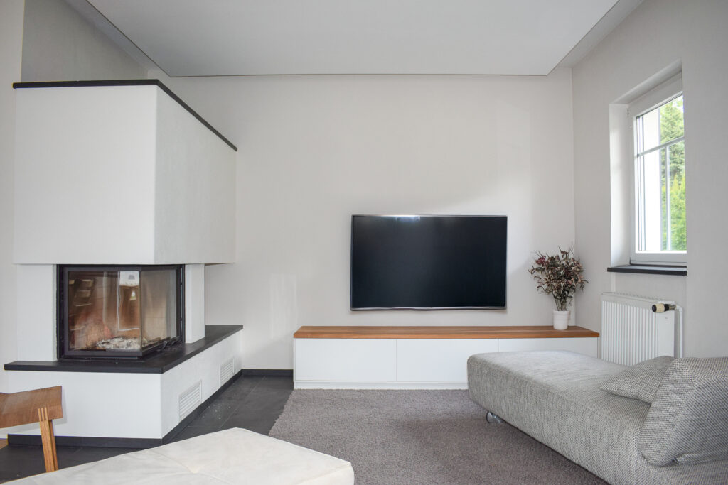 Modernes Wohnzimmer mit einem an der Wand montierten Fernseher, einem eingebauten Kamin, einem grauen Sofa, einem weißen niedrigen Schrank, einem kleinen Tisch, einem Holzstuhl und einem Fenster mit einer Blumenvase auf der Fensterbank.