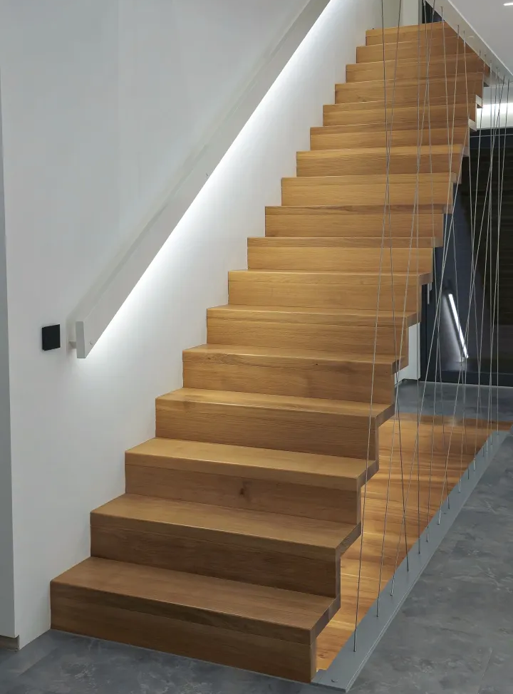 Eine moderne Innentreppe aus hellem Holz mit breiten Stufen, eingefasst von dünnen vertikalen Metallkabeln und beleuchtet von einem weißen LED-Lichtband entlang der Wand.