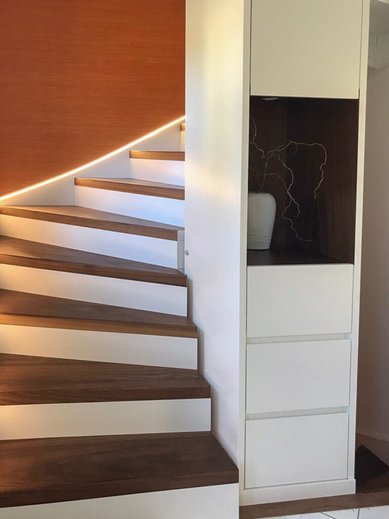 Moderne Holztreppe mit weißen Setzstufen und einem Band mit warmer LED-Beleuchtung entlang der geschwungenen Wand. Neben der Treppe steht ein weißer Einbauschrank mit einer dekorativen Vase und Wandkunst in einem eingelassenen Regal.
