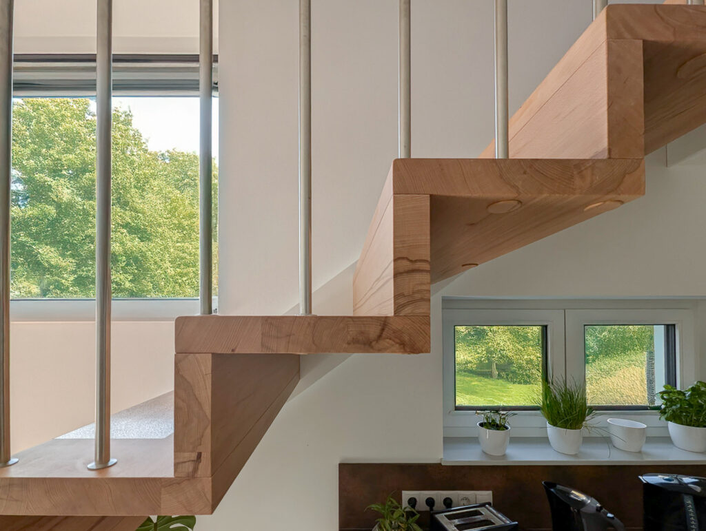 Eine moderne Holztreppe mit Metallgeländer, die neben großen Fenstern steht, die viel Tageslicht hereinlassen. Auf der Fensterbank darunter stehen Topfpflanzen, während draußen grüne Bäume zu sehen sind.