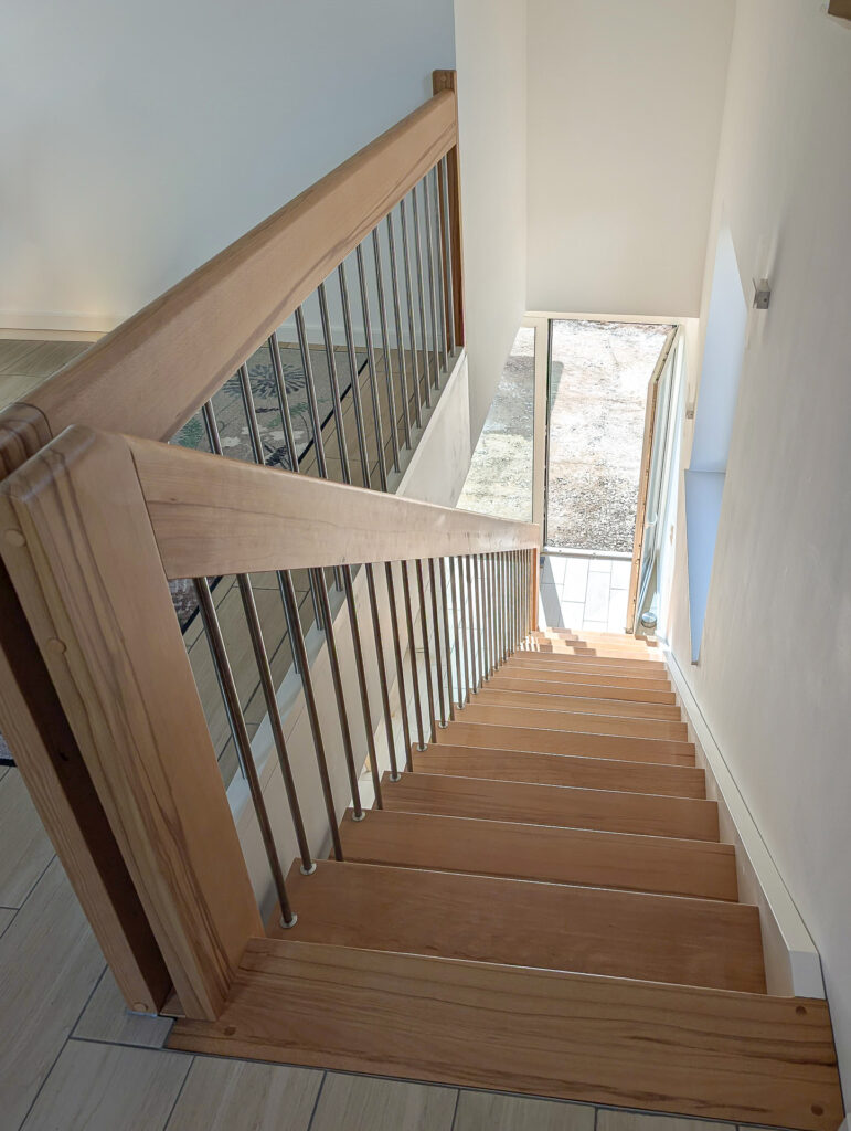 Blick vom oberen Ende einer Holztreppe mit Metallgeländerstäben, die zu einer Glastür hinunterführt, die zu einem Außenbereich mit einfallendem Sonnenlicht führt.