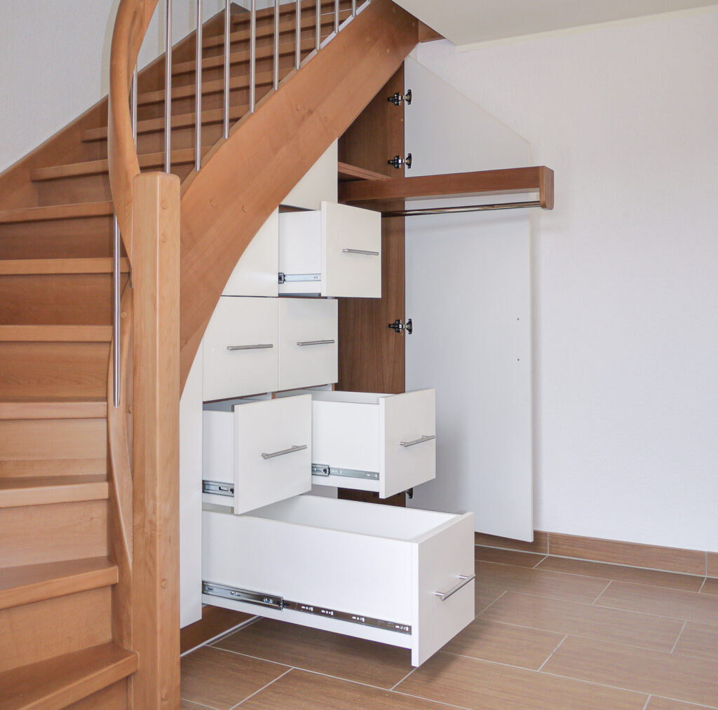 Eine Holztreppe mit eingebauten weißen Schubladen und Schränken darunter, von denen einige offen sind, um den Platz optimal zu nutzen. Der Boden ist hellbraun gefliest, die Wände sind weiß.