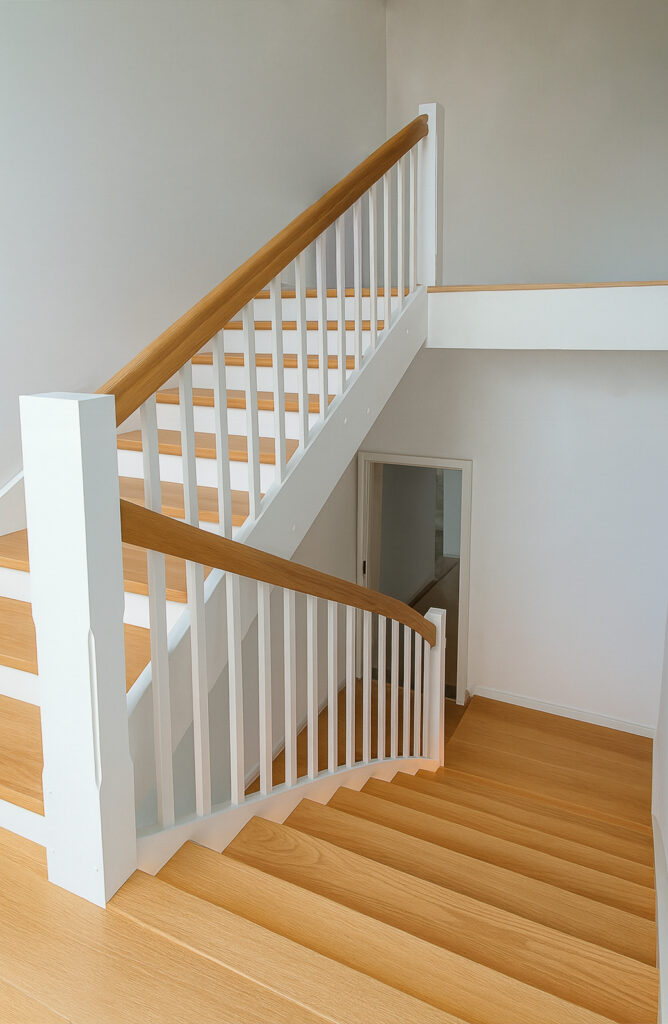 Eine moderne Innentreppe mit hellen Holzstufen, weißen Setzstufen und weißem Geländer mit Holzhandlauf, die zu einem kleinen Treppenabsatz und einer weißen Tür im unteren Stockwerk führt.